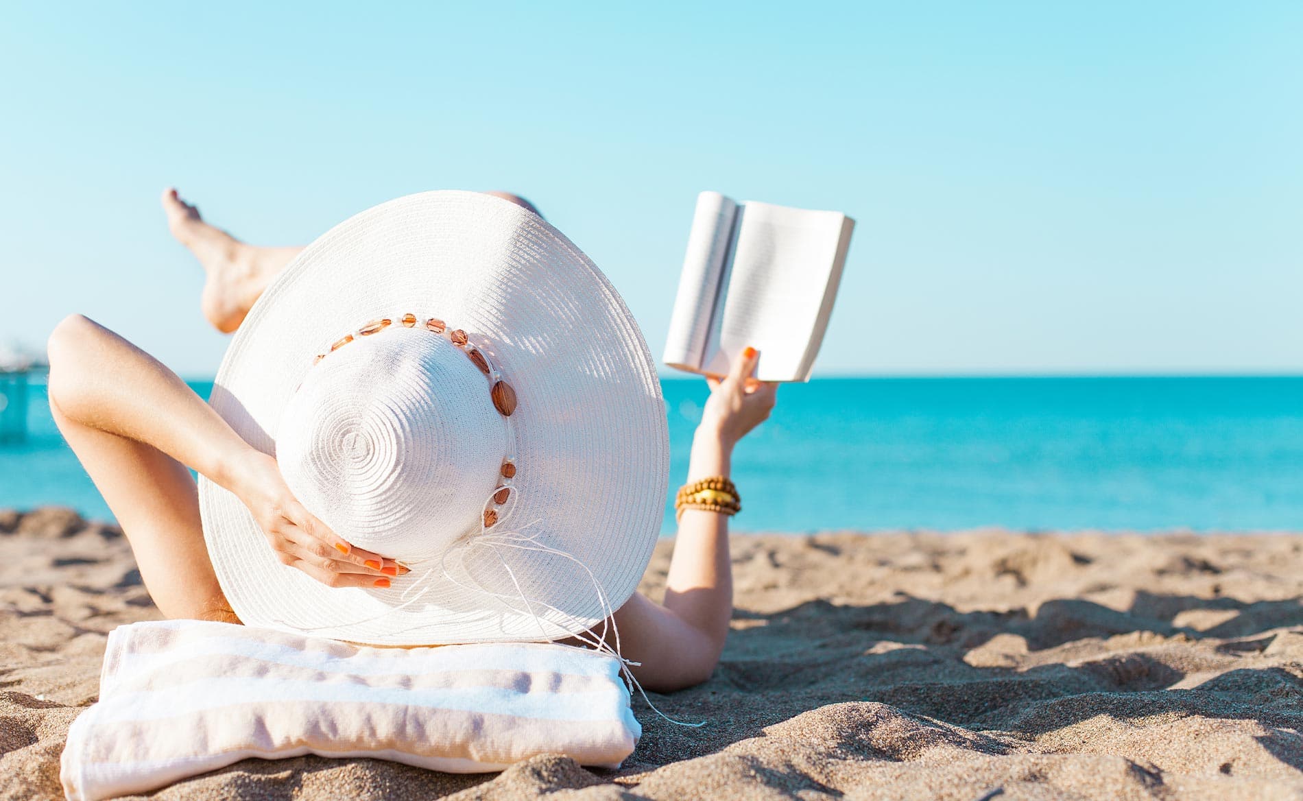 A person lounging on the beach in a large white hat, reading a book by the ocean.