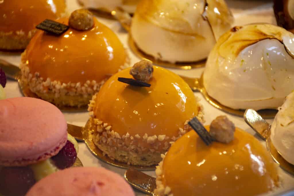 An assortment of colorful pastries, including orange-glazed dome cakes and pink macarons, displayed in a bakery.