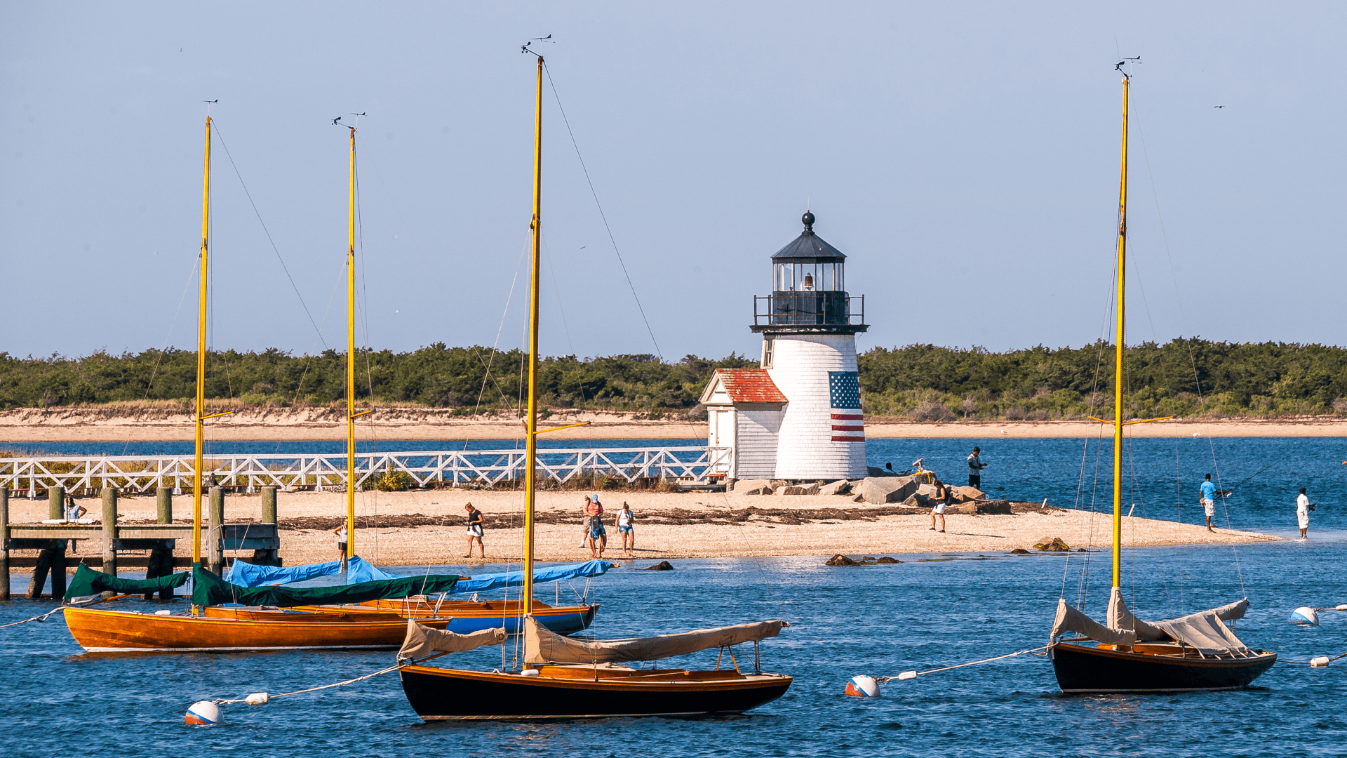 A lighthouse stands on a sandy shore, surrounded by boats in the water and people walking nearby. A lighthouse stands on a sandy shore, surrounded by boats in the water and people walking nearby.