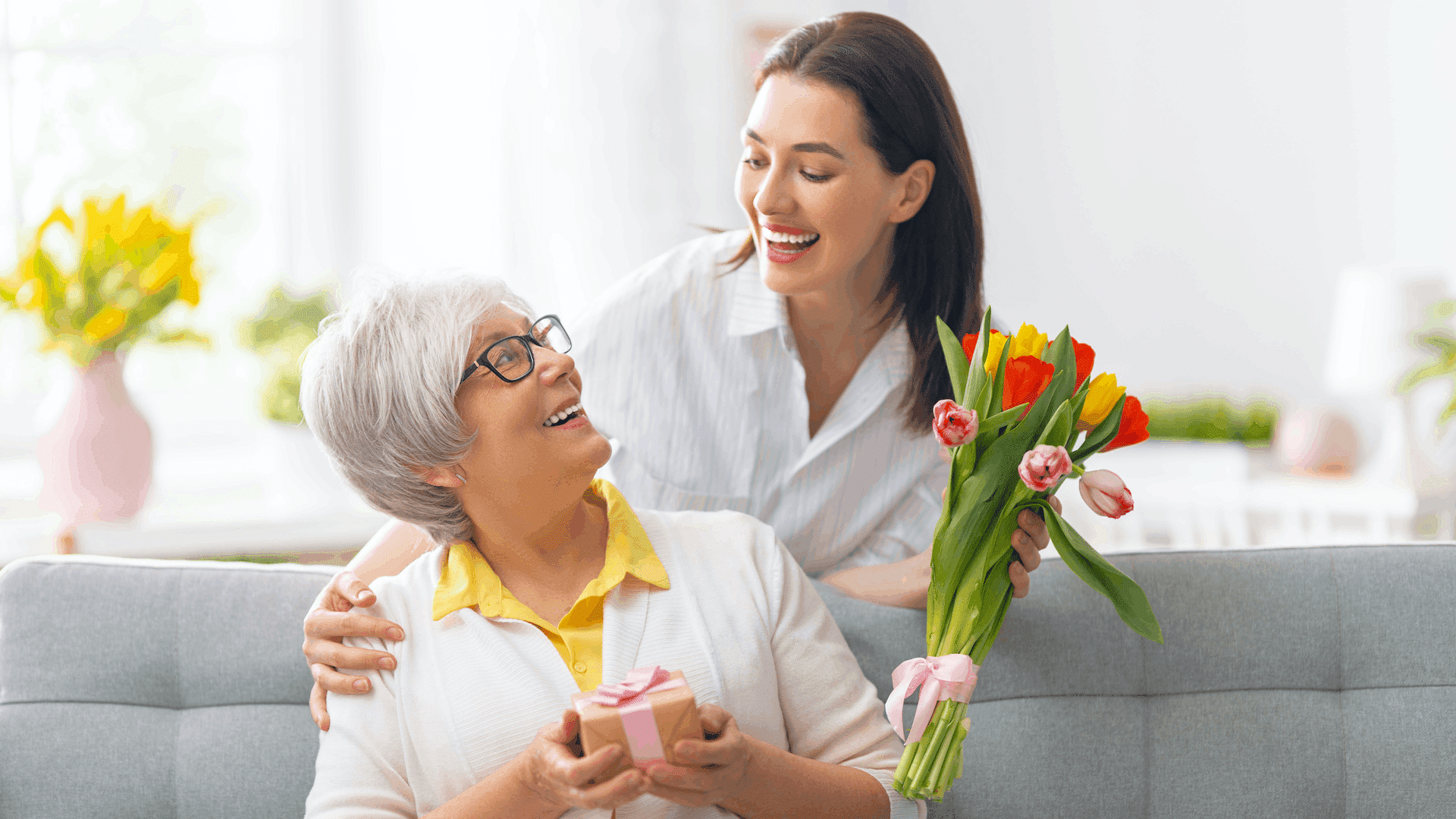 A woman surprises her smiling mother with a gift and flowers in a bright living room.