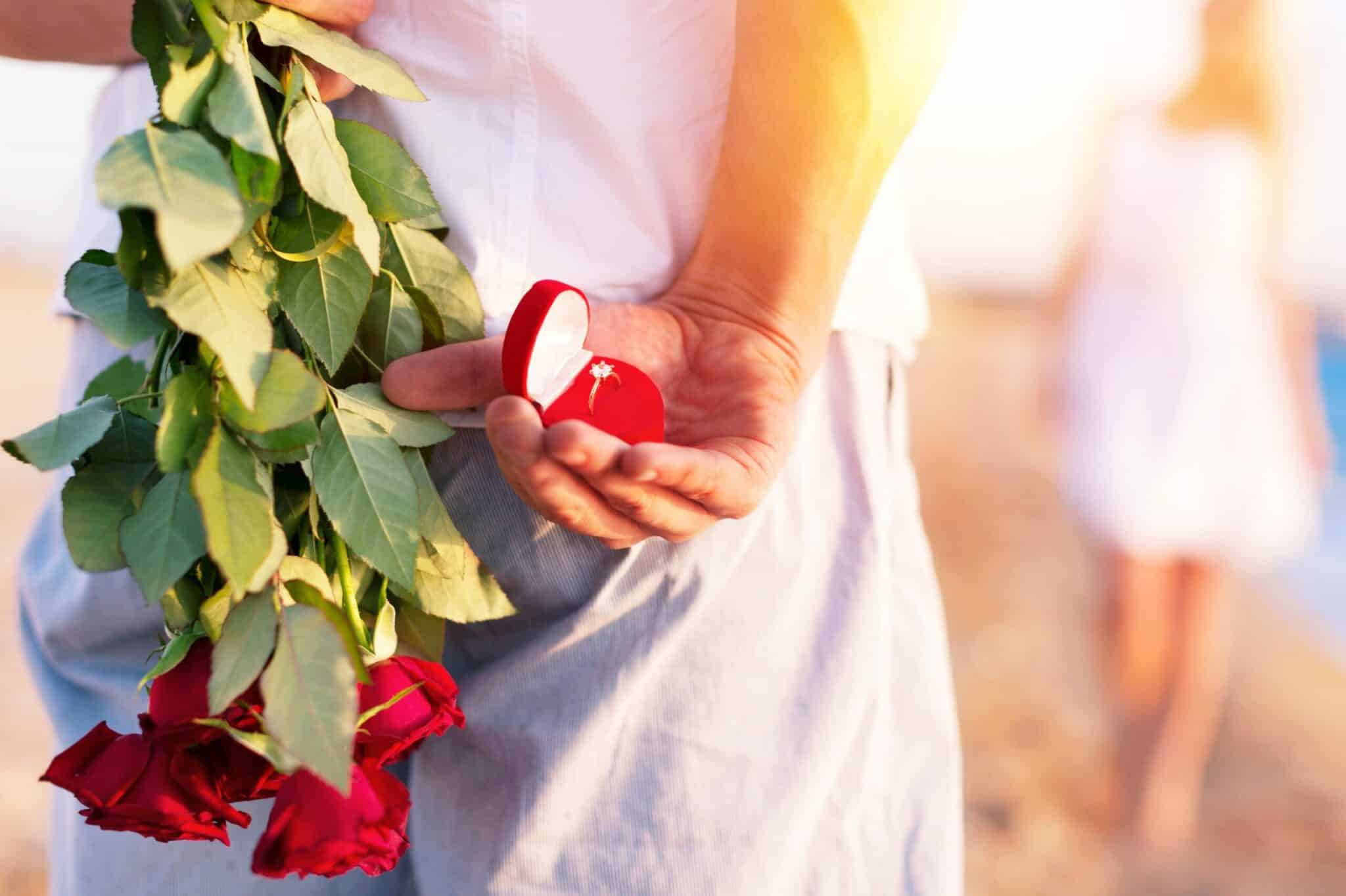 A person holding a bouquet of red roses and a ring box behind their back stands near a woman walking away.
