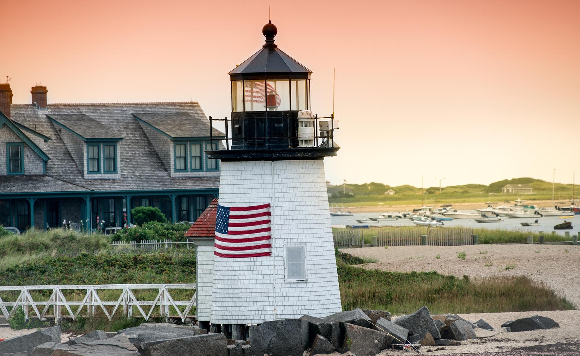A white lighthouse with an American flag stands beside a beach at sunset, with a house and boats in the background.