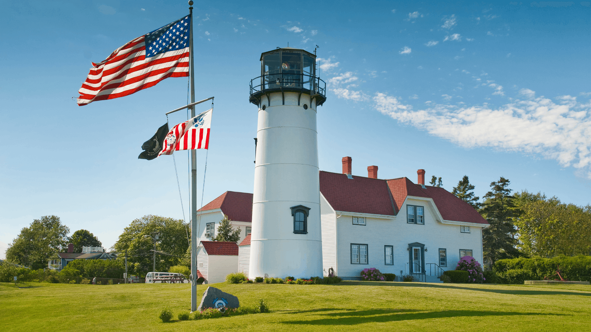 A white lighthouse with a red-roofed building and flags in front, set against a clear blue sky.