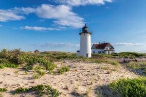 A white lighthouse stands beside a sandy beach and greenery under a blue sky.