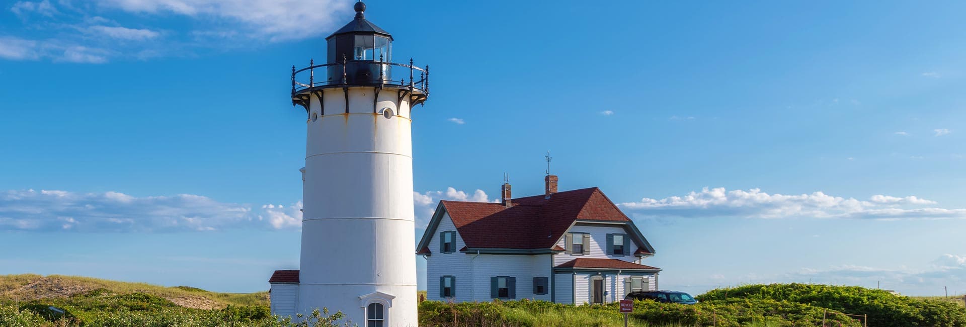 A white lighthouse stands beside a house against a clear blue sky with scattered clouds.