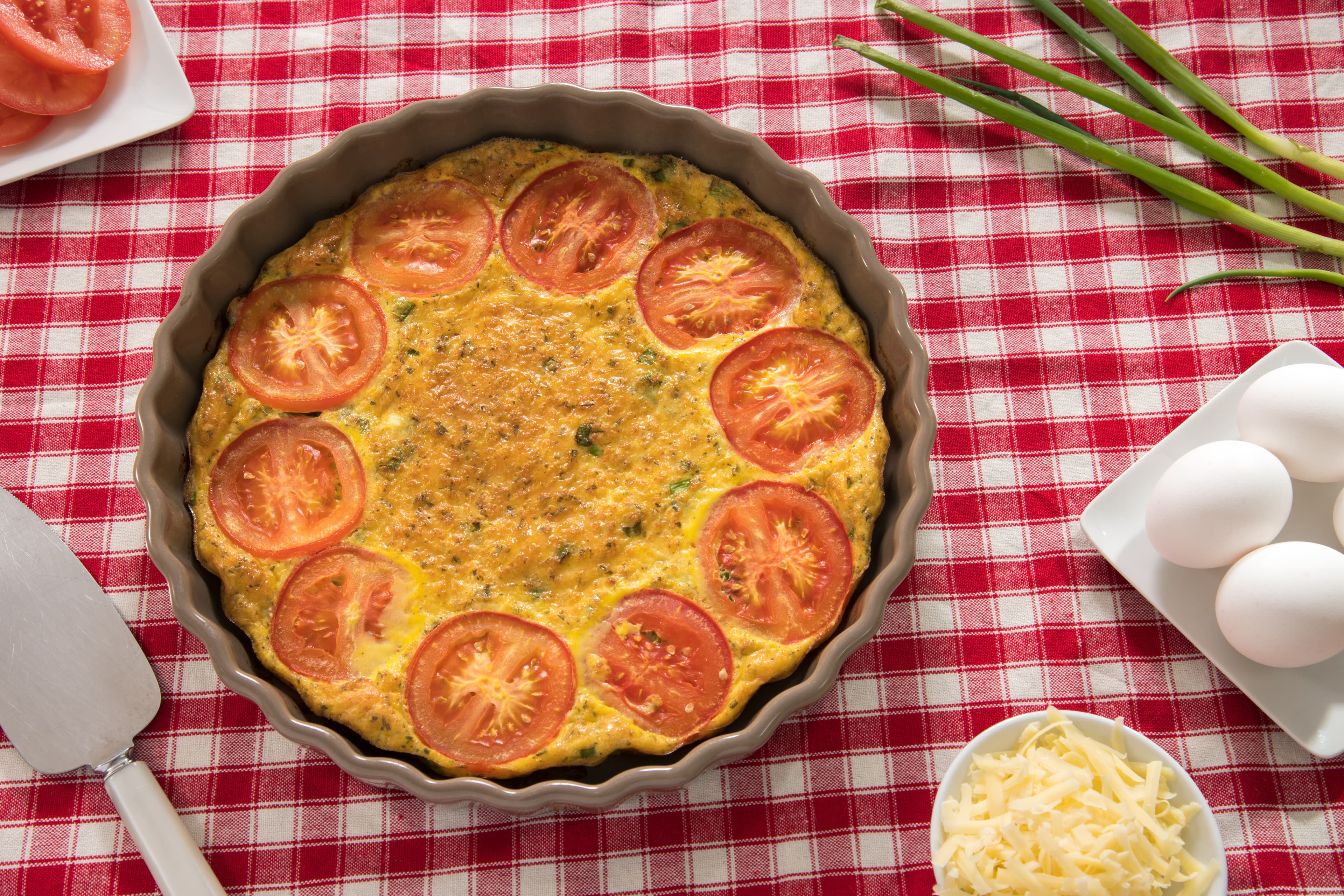 A baked frittata topped with tomato slices, surrounded by fresh vegetables and ingredients on a checkered tablecloth.