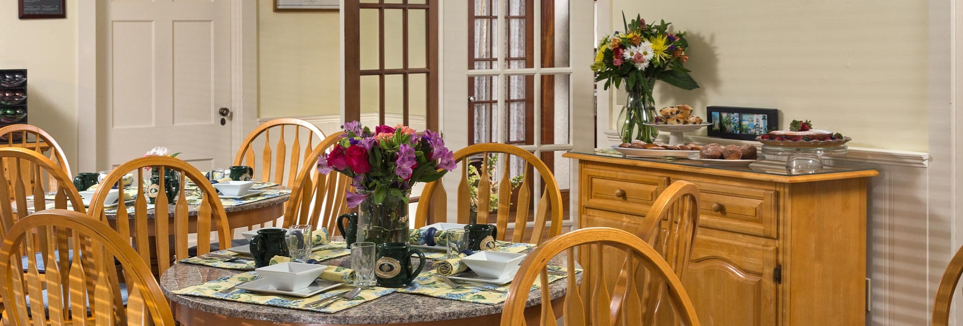 A cozy dining area with a wooden table set for a meal, featuring flowers and a side buffet.