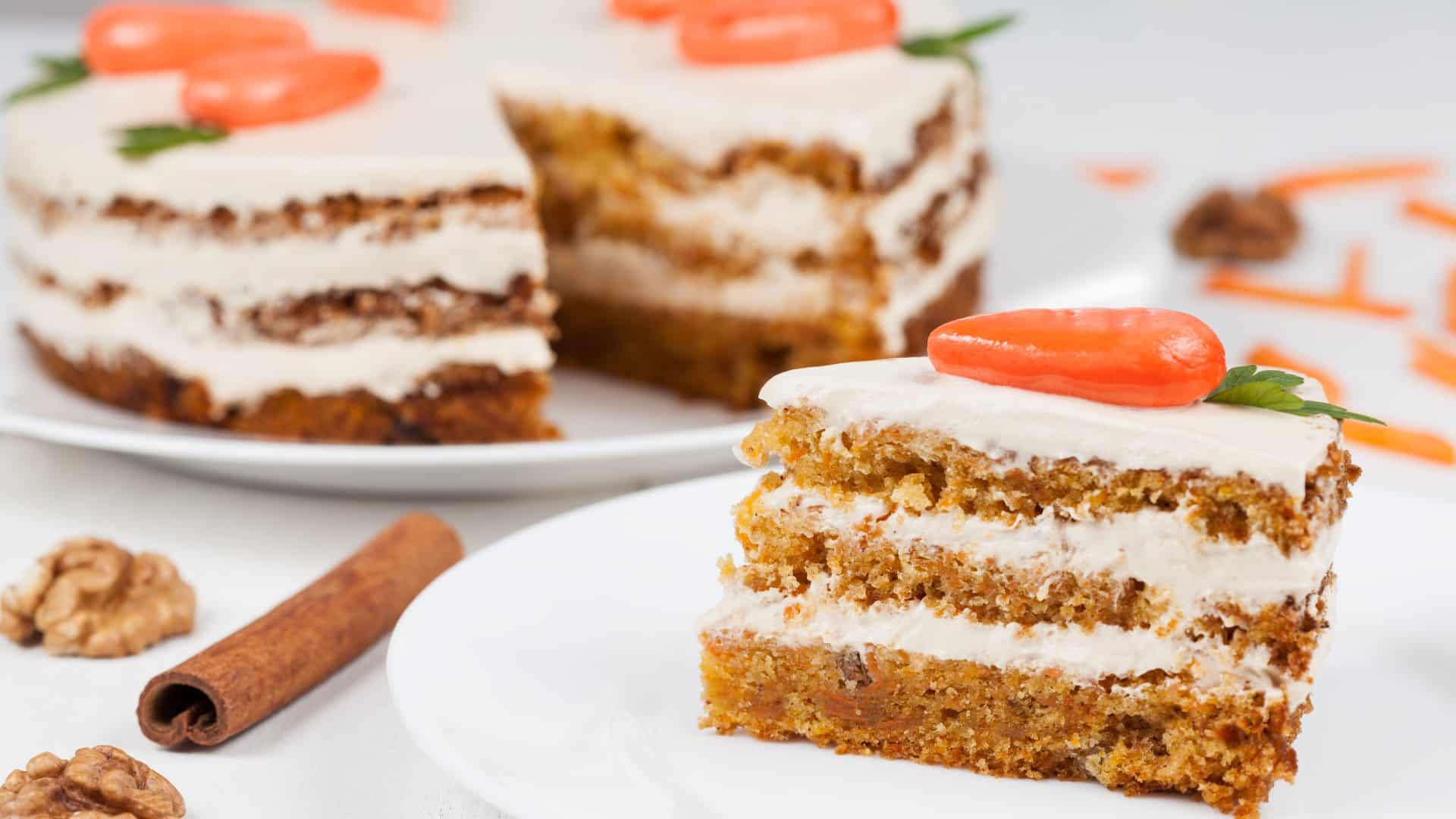 A slice of layered carrot cake topped with cream cheese frosting and a decorative carrot, with the whole cake partially visible in the background. A slice of layered carrot cake topped with cream cheese frosting and a decorative carrot, with the whole cake partially visible in the background.