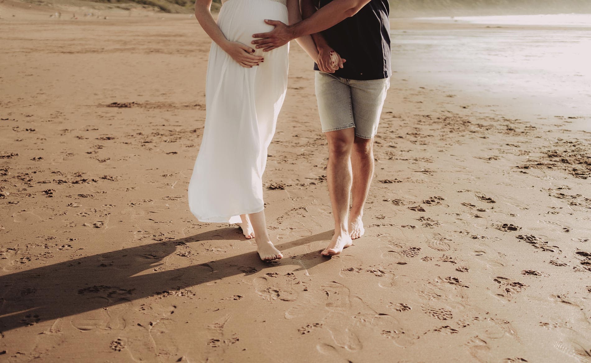 A couple stands barefoot on a beach, with the woman cradling her baby bump.