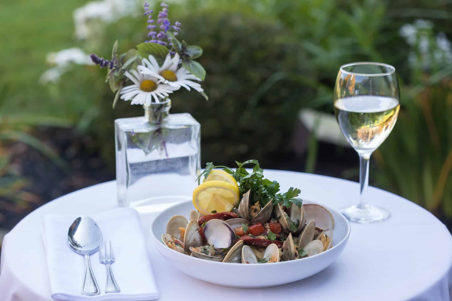 A bowl of clams with herbs and lemon, accompanied by a glass of white wine and a flower vase, on a table outdoors.