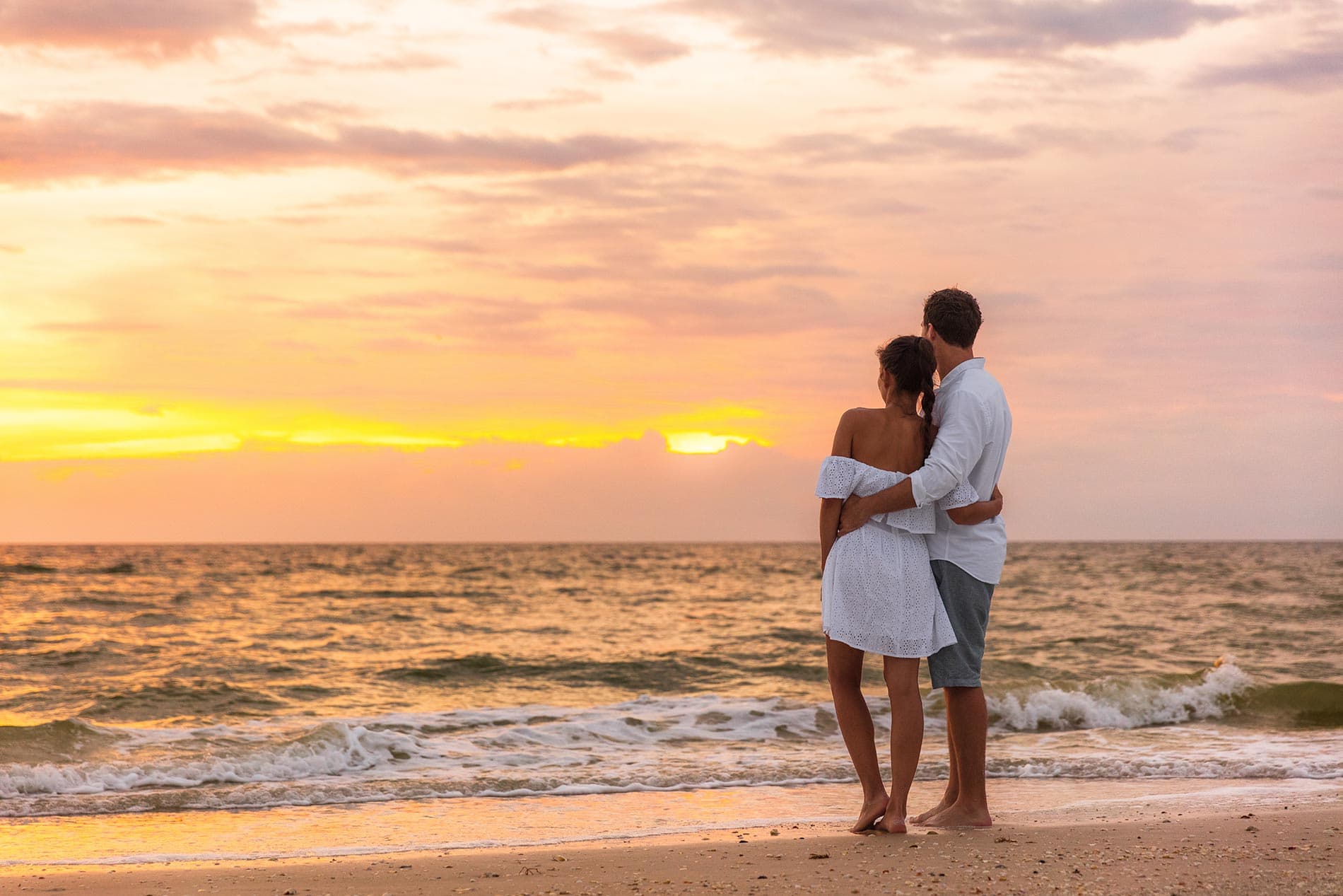 A couple embraces on the beach at sunset, gazing at the ocean.