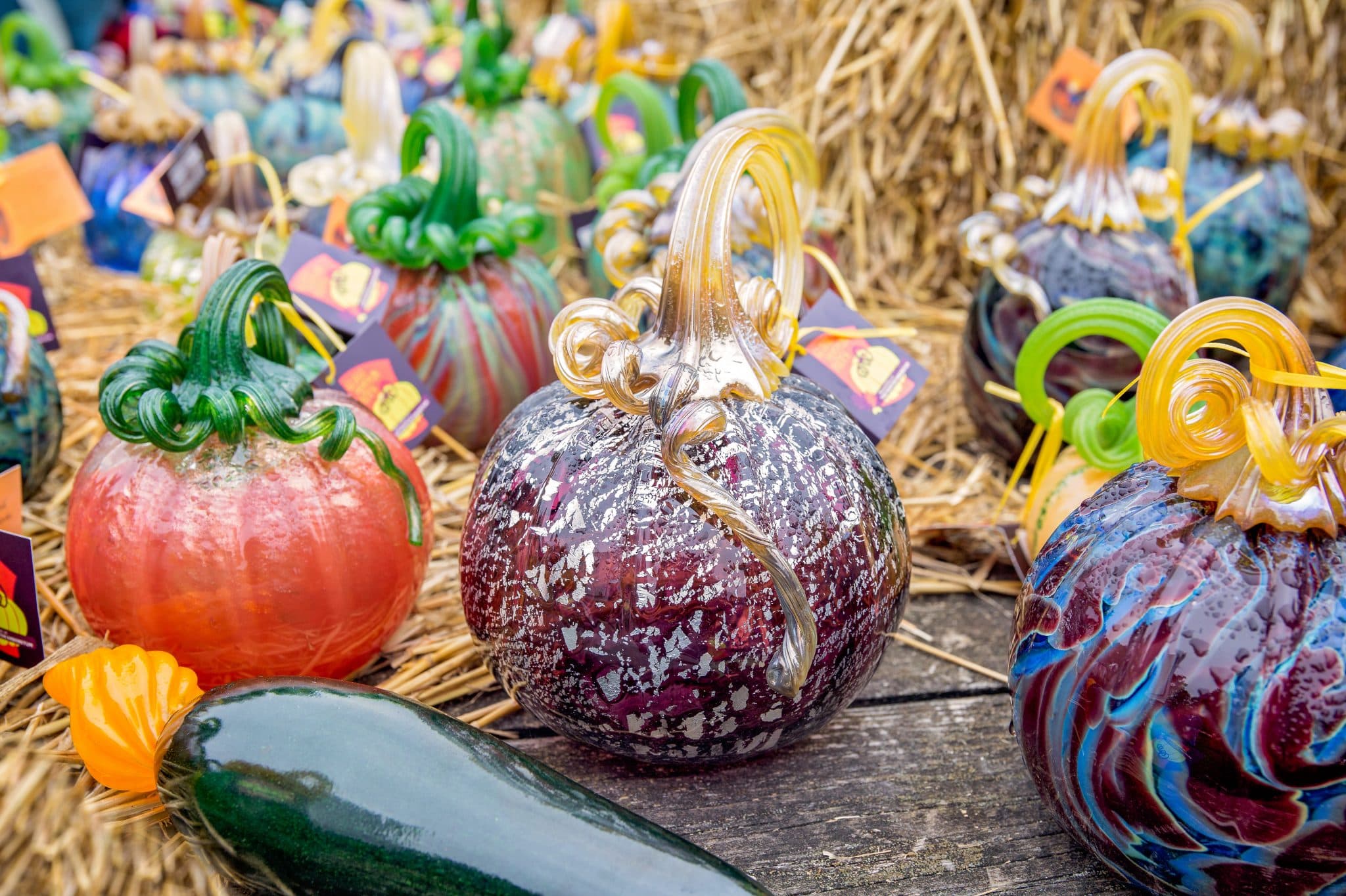 A collection of colorful glass pumpkins and gourds displayed on wooden and straw surfaces.