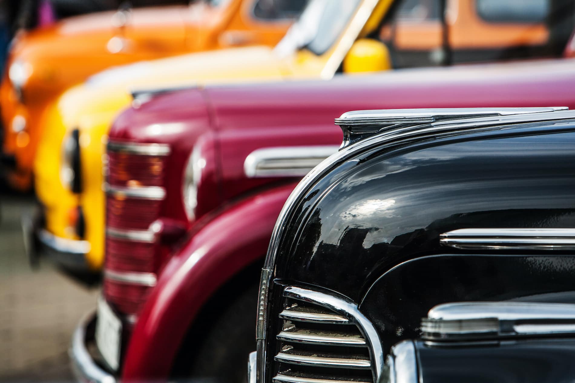 A row of vintage cars in various colors, featuring shiny hoods and distinctive chrome details.