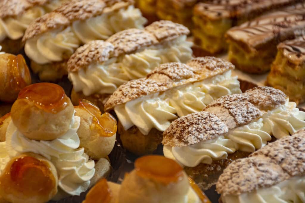 A variety of cream-filled pastries, including éclairs and cream puffs, adorned with whipped cream and powdered sugar.