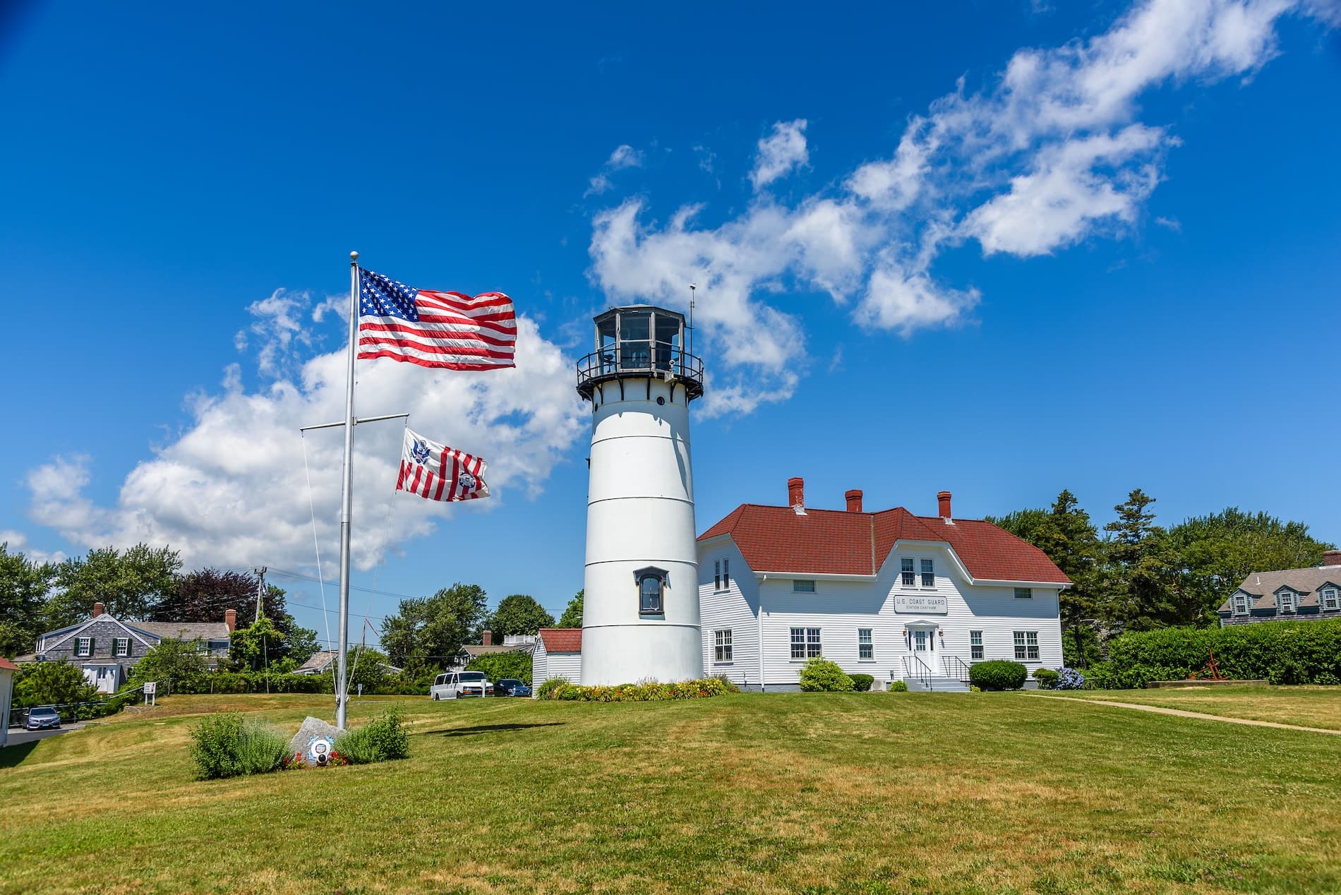 A white lighthouse stands beside American flags against a bright blue sky.