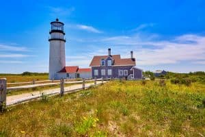 A lighthouse stands beside a historic house in a grassy landscape under a clear blue sky.
