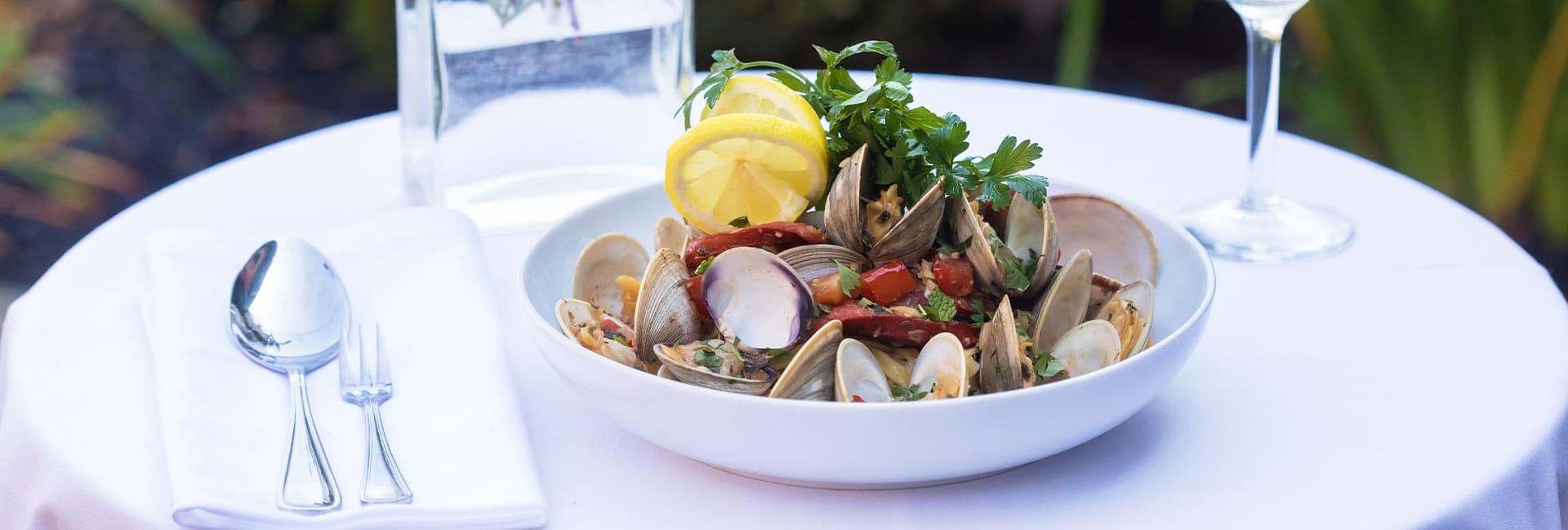 A bowl of clams garnished with lemon and herbs on a white tablecloth, accompanied by cutlery and a glass of water.