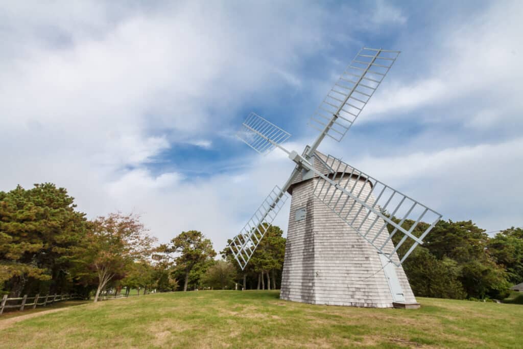 A weathered wooden windmill stands on a grassy hill under a cloudy sky.