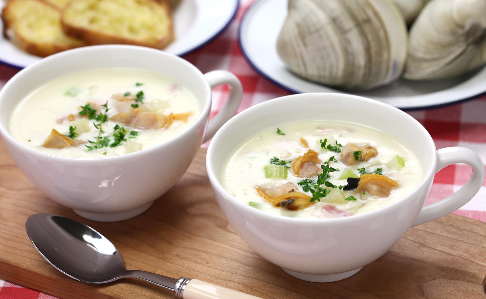 Two bowls of clam chowder garnished with parsley on a wooden board, accompanied by toast and clams in the background.