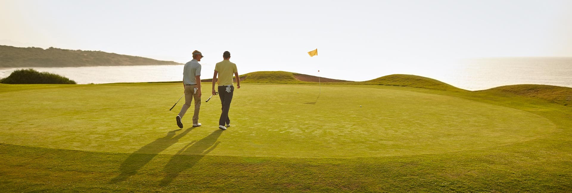 Two golfers walk together on a sunlit green overlooking the ocean.