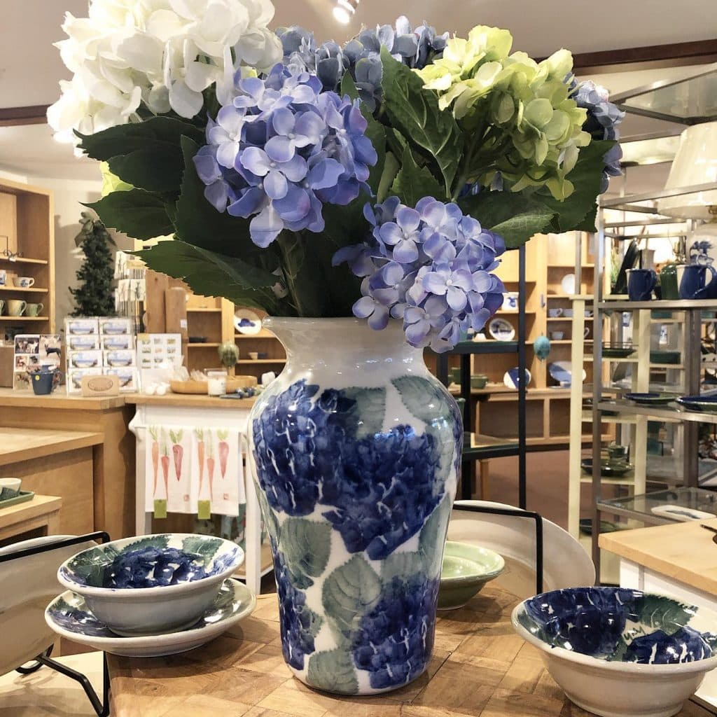 A decorative vase with blue and white floral patterns holds hydrangeas in various shades of blue and green, surrounded by ceramic bowls.