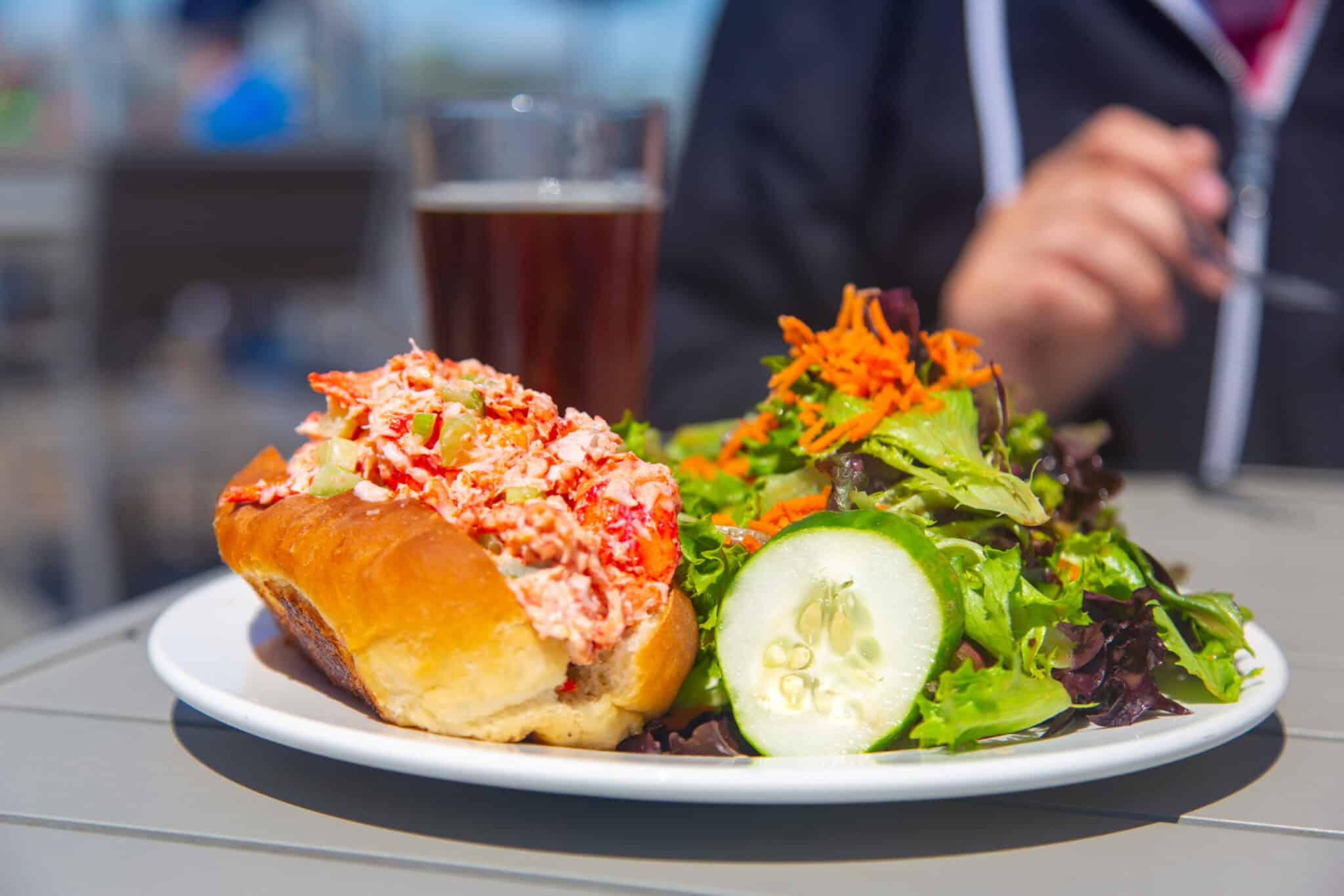 A lobster roll with a side salad and a glass of dark beer on a table.