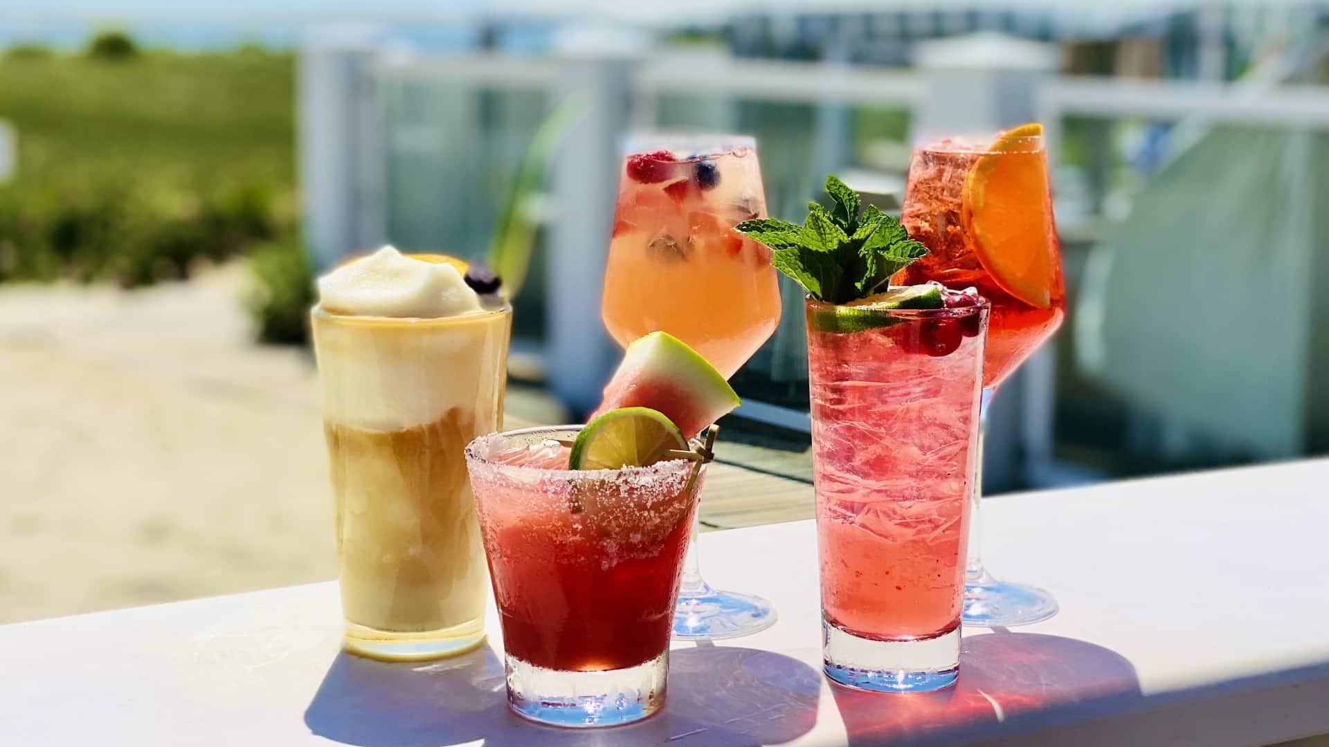 Four colorful cocktails with garnishes are displayed on a bar against a beach backdrop.