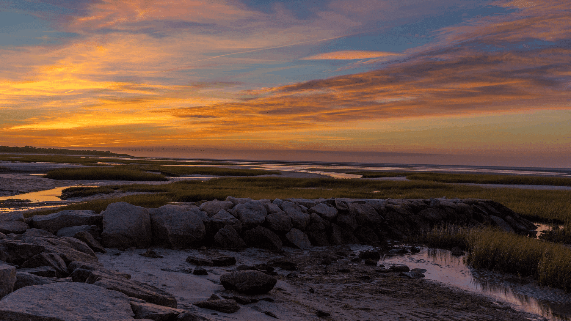 A serene sunset over a coastal marsh with rocky shorelines and gentle grasses.