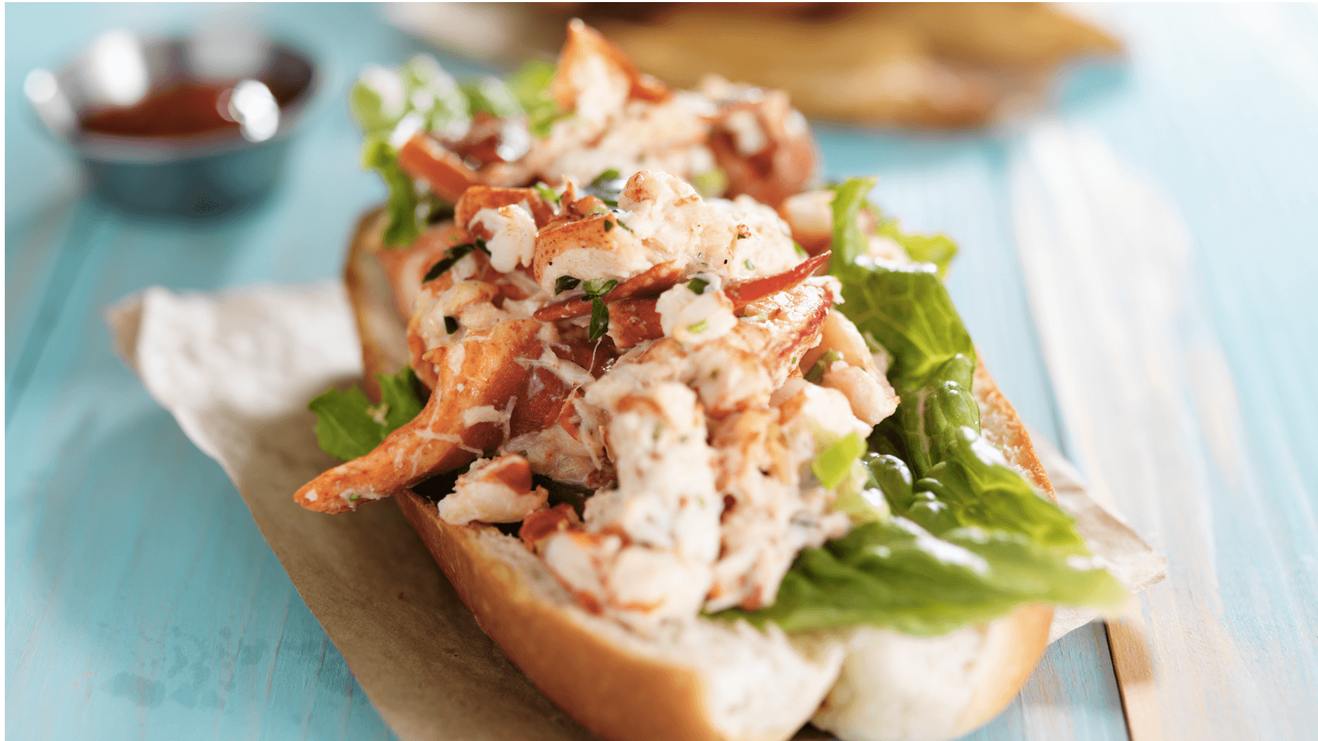 Lobster roll topped with lettuce and herbs on a light wooden table.
