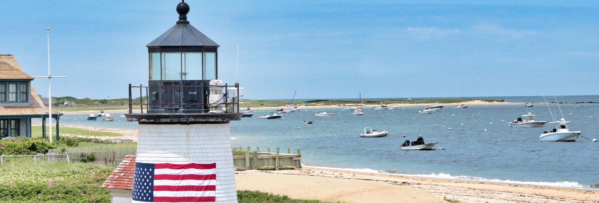 A lighthouse with an American flag overlooks a beach with boats in the water.