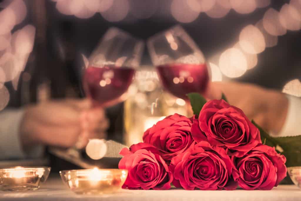 A romantic dinner setting with glasses of wine clinking and a bouquet of red roses in the foreground.