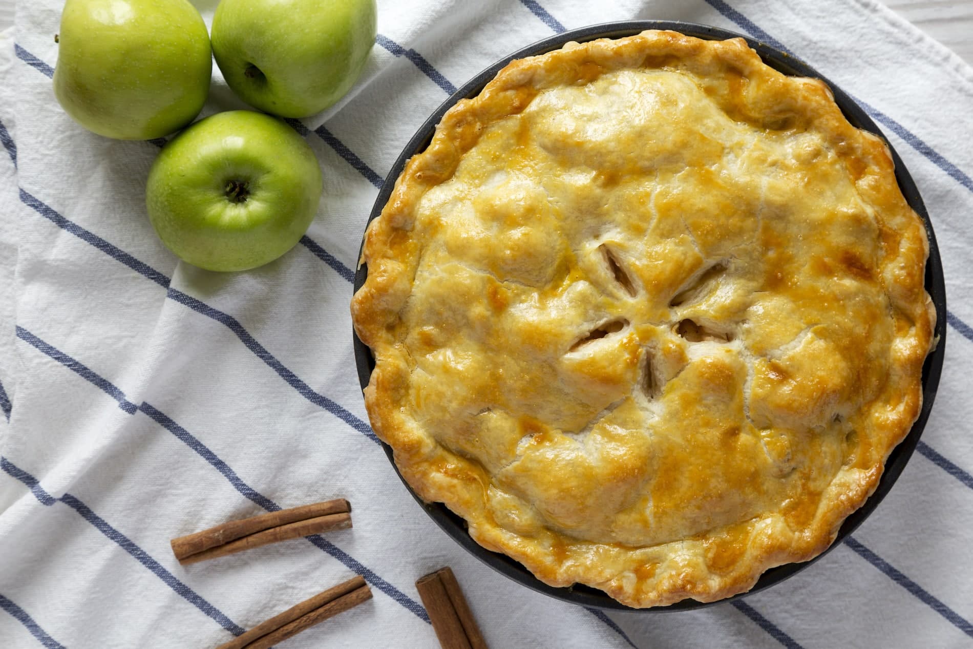 A golden-brown apple pie surrounded by green apples and cinnamon sticks on a striped cloth.