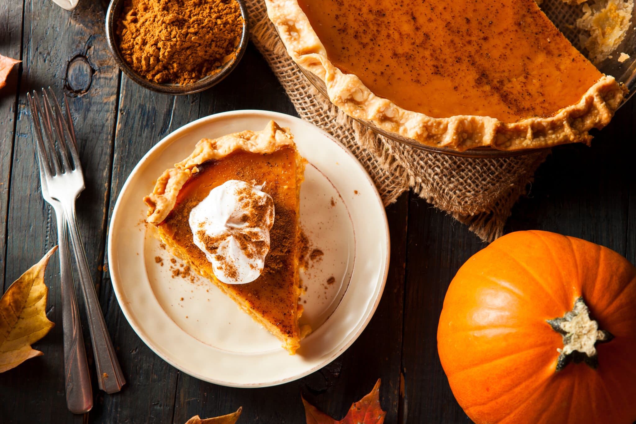 A slice of pumpkin pie topped with whipped cream on a white plate, surrounded by a whole pumpkin, autumn leaves, and a bowl of pumpkin spice. A slice of pumpkin pie topped with whipped cream on a white plate, surrounded by a whole pumpkin, autumn leaves, and a bowl of pumpkin spice.