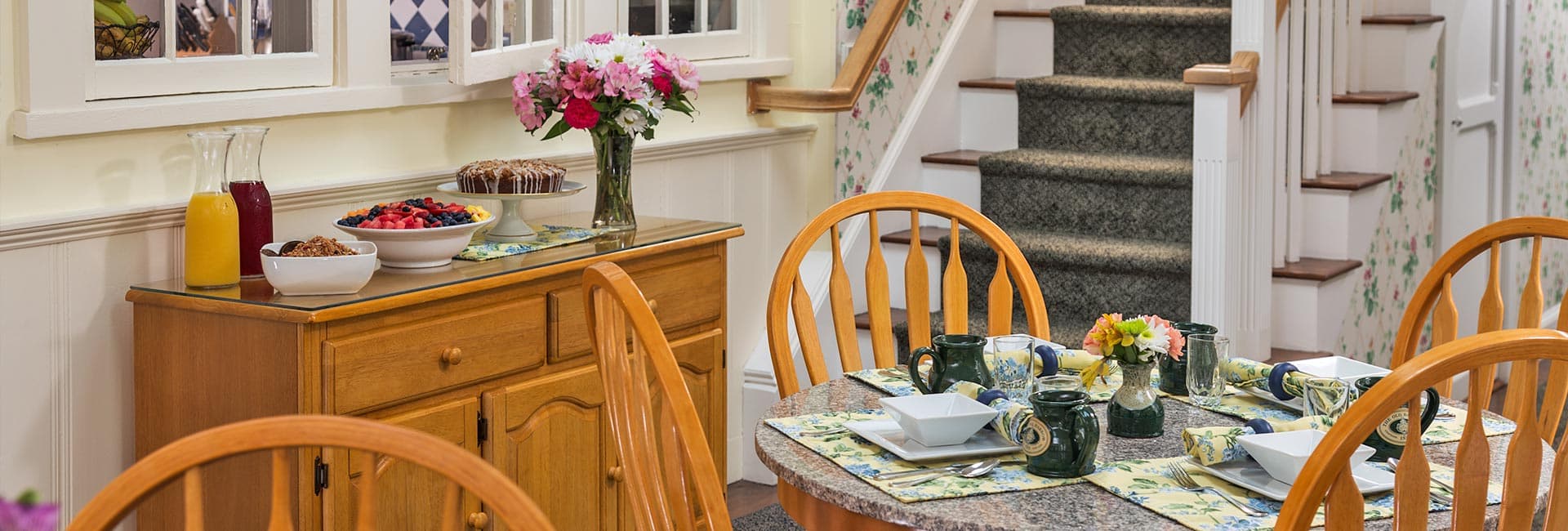 A cozy dining area featuring a wooden table set with dishes and surrounded by chairs, with a sideboard displaying food and flowers.