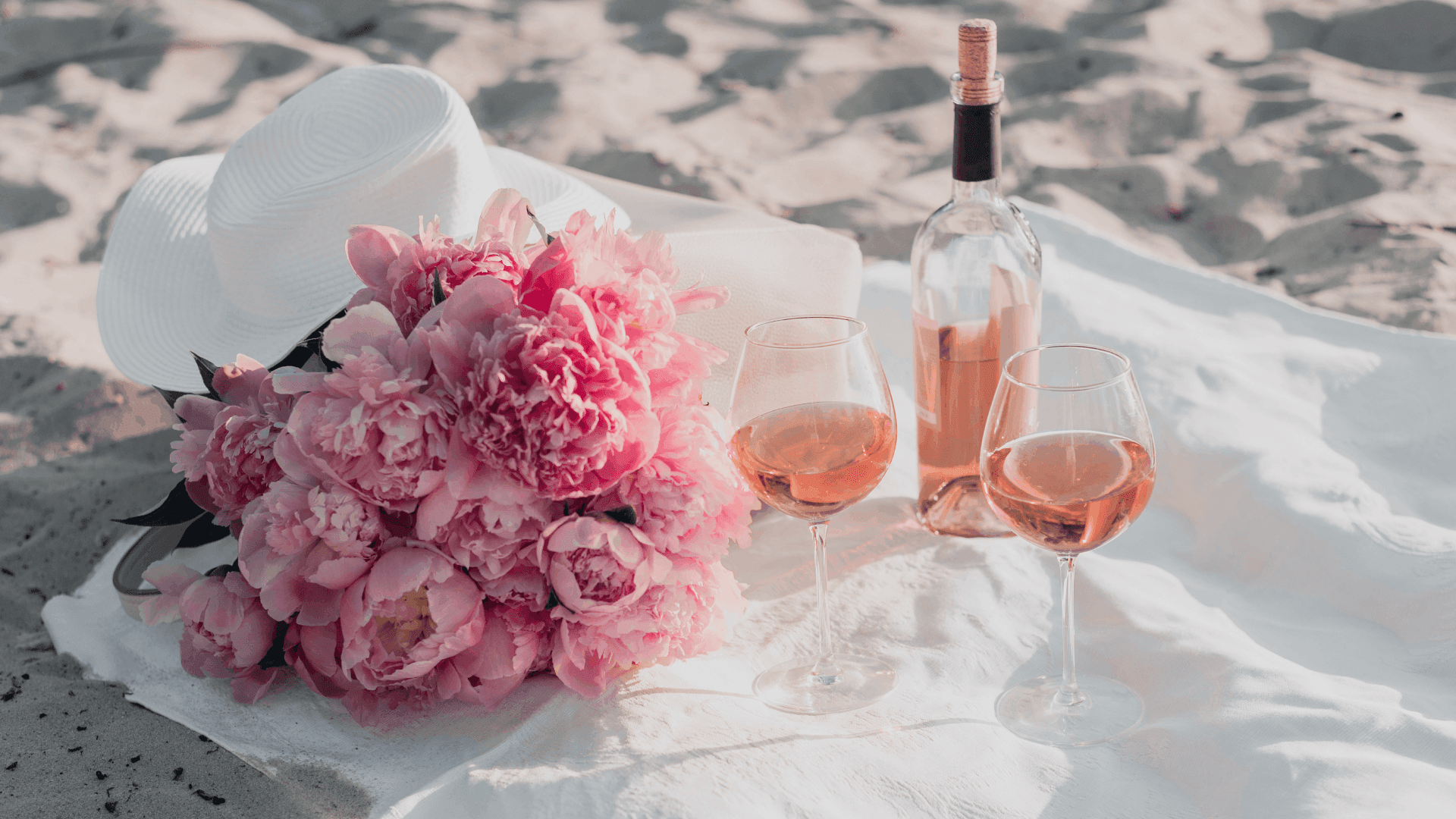 A bouquet of pink peonies, two glasses of rosé, and a bottle of wine are arranged on a white cloth in a sandy setting.