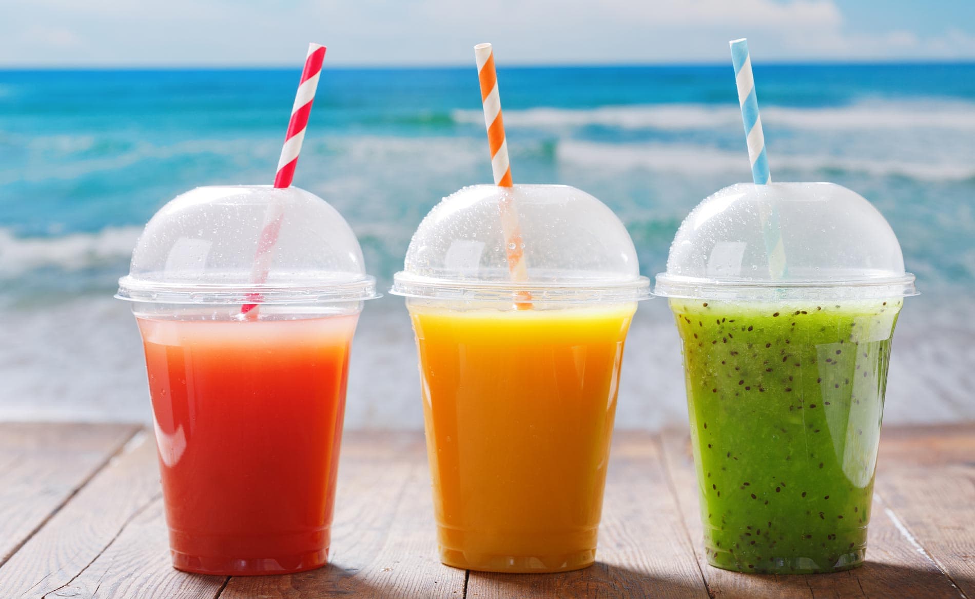 Three colorful fruit drinks with straws sit on a wooden surface, with the ocean visible in the background.
