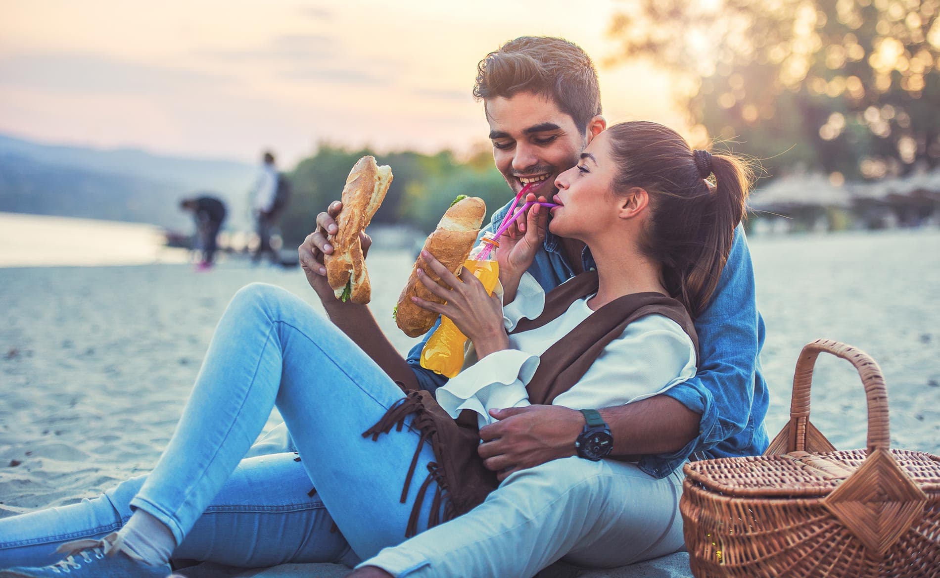 A couple enjoys sandwiches while sitting on the beach at sunset.