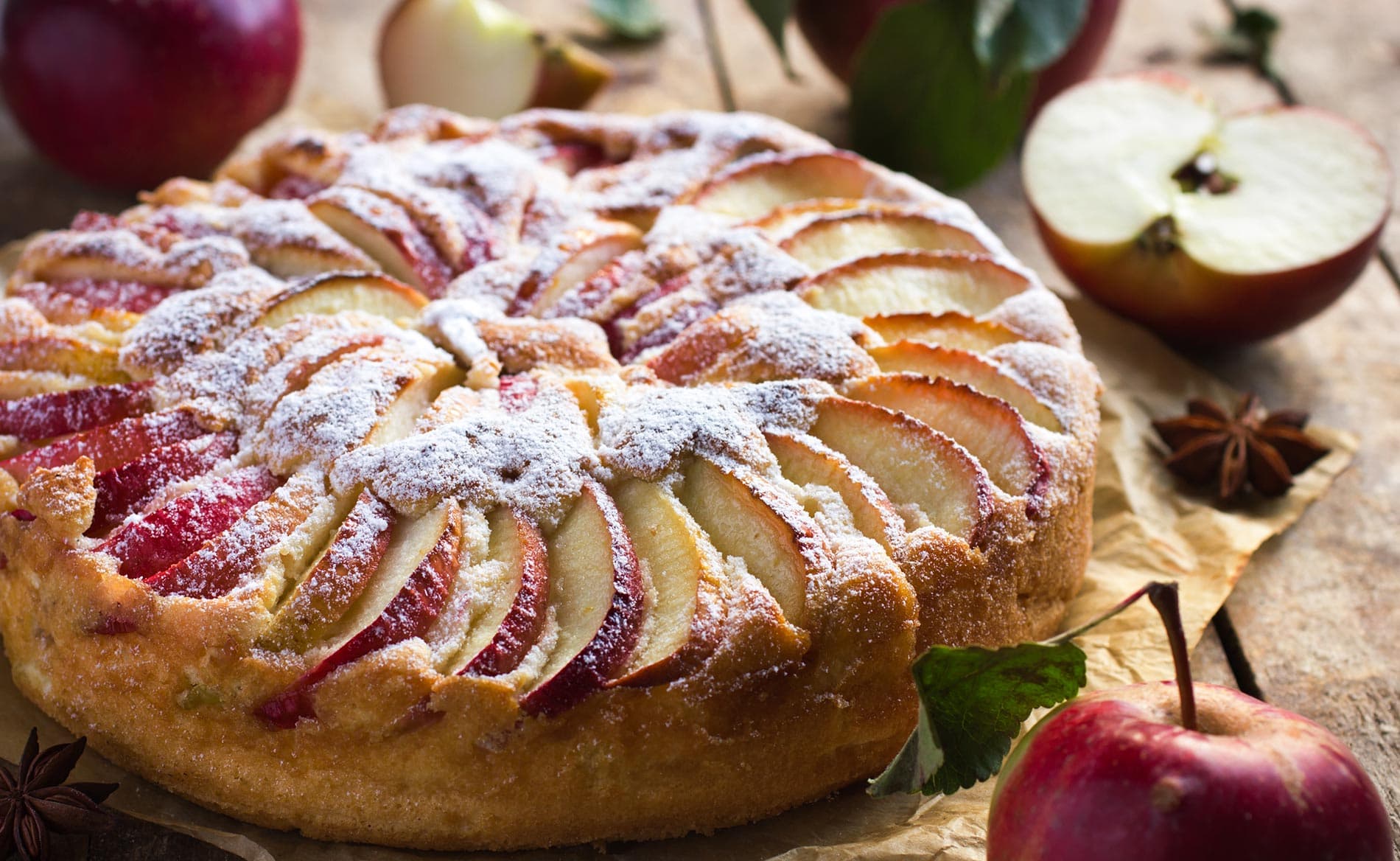 A freshly baked apple tart dusted with powdered sugar, surrounded by apples and greenery on a wooden surface.