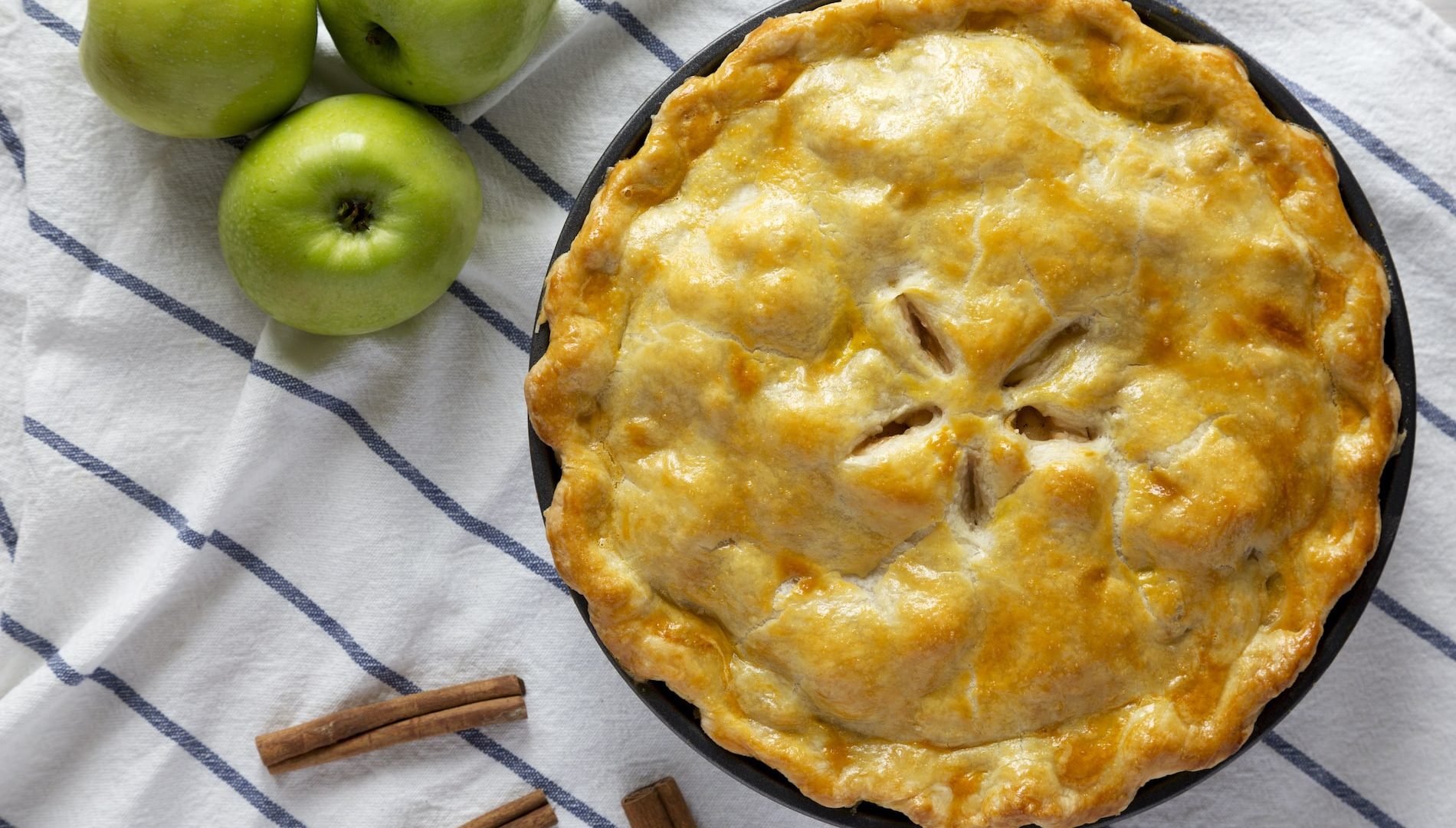 Homemade apple pie on cloth with three green apples and a few cinnamon sticks , top view. Flat lay, overhead, from above.