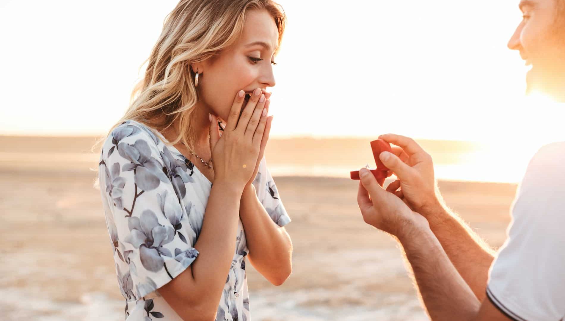 Photo of smiling man making proposal to his amazed woman with ring in gift box while walking on sunny beach