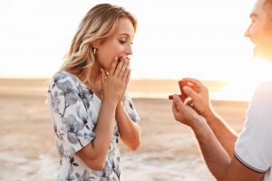 Photo of smiling man making proposal to his amazed woman with ring in gift box while walking on sunny beach