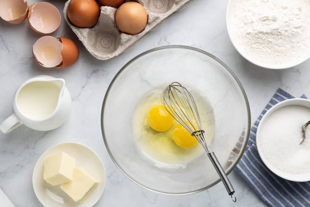 baking cake ingredients (eggs, flour, sugar, butter and milk) on white table, top view