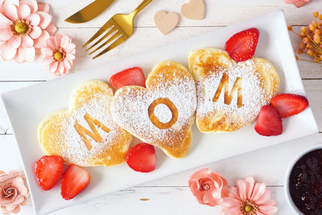Heart shaped pancakes with MOM letters. Mothers Day breakfast concept. Top view table scene with a white wood background.