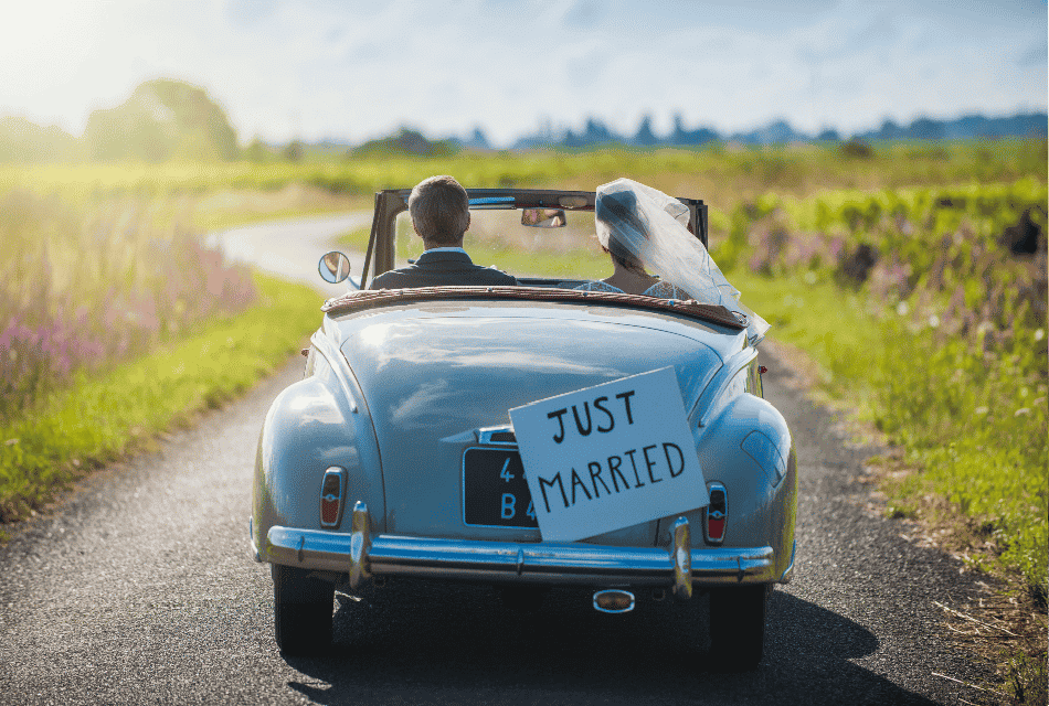 A married couple in a retro light blue car with sign just married driving on a road surrounded by greenery