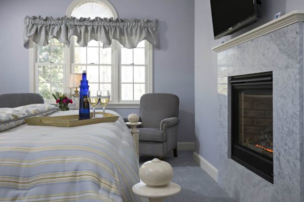 Light BLue guest room with triple window, upholstered seating, bed topped with wine, tv and gas fireplace