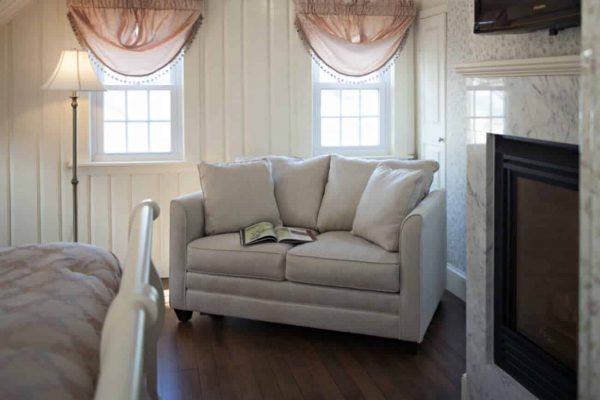 Guest room with creamy paneled walls, sunny windows, wood floors and cozy love seat next to a gas fireplace