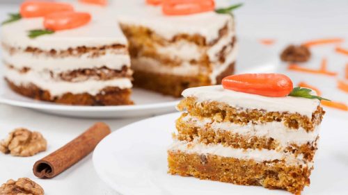 Slice of three layer carrot cake with cream cheese frosting and decorative carrot on top