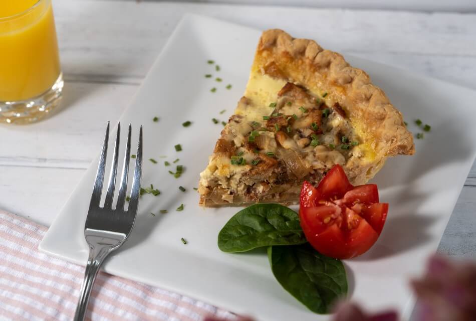 A slice of breakfast quiche on a white plate with tomato, basil leaves and a silver fork