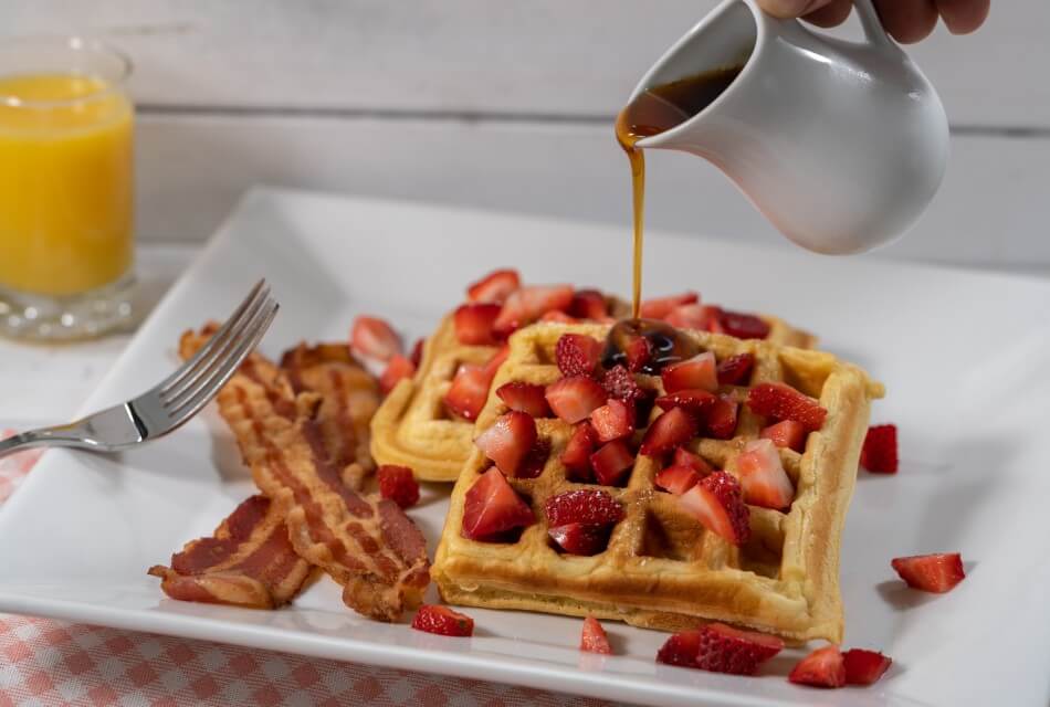 White plate of waffles, berries and bacon with syrup being poured
