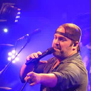 Male, lead singer of a band wearing a backwards baseball cap, singing into a microphone and pointing to the audience